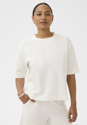 Femme aux cheveux attachés portant un haut blanc à manches courtes et un pantalon, debout avec une main dans la poche devant un fond uni.