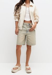 Chaqueta beige clara, top corto blanco, shorts de mezclilla gastada y sandalias de plataforma beige. Texturas suaves y ajuste relajado en todo el conjunto.