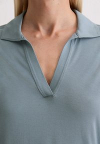 Jasnoniebieska koszulka polo z dekoltem w serek. Wykonana z miękkiego materiału, z wyraźnie zarysowanym kołnierzykiem i gładką teksturą. Brak widocznych wzorów.