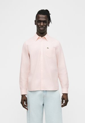 Hombre de piel oscura y rastas que lleva una camisa rosa claro abotonada y vaqueros azul claro, de pie frente a un fondo blanco liso.