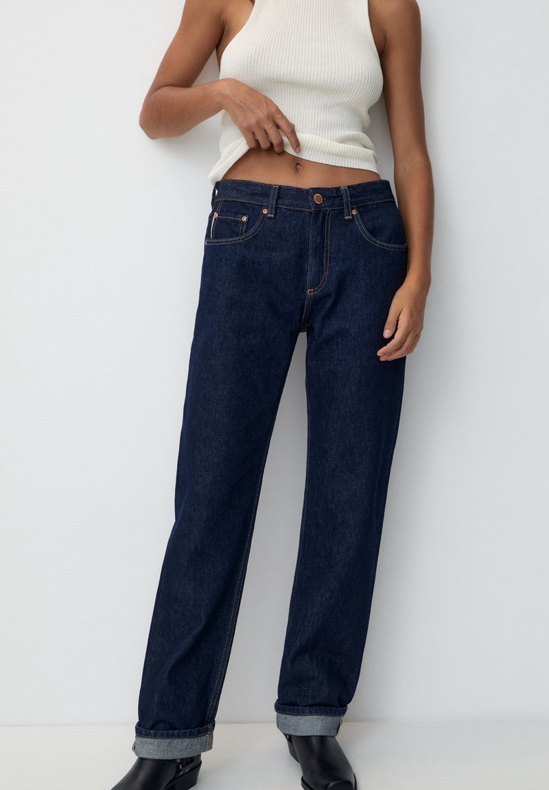 Femme portant un jean droit bleu foncé avec des revers, un haut blanc côtelé sans manches et des bottines noires à talons.