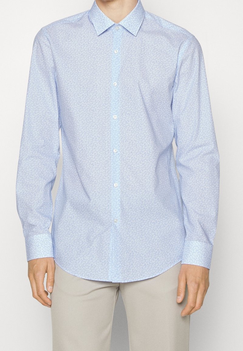 Homme portant une chemise à manches longues bleue claire à motifs, boutonnée, avec un pantalon beige, debout devant un fond uni.