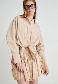 Licht geruite blouse in beige met een geknoopte voorkant; gecombineerd met een korte, geplooide blushrok met ruwe randen en een elastische tailleband.
