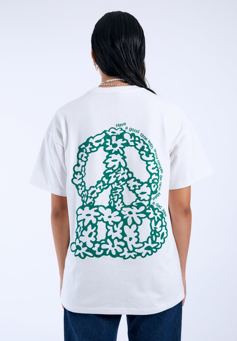 Bílý bavlněný tričko s velkým zeleným květinovým grafickým motivem mírového znamení na zádech, obklopeným zakřiveným textem. Krátké rukávy, uvolněný střih.