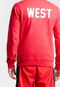 Sweat-shirt rouge avec l'inscription "WEST" en blanc au dos, doté de poignets côtelés et d'un col rond classique. Texture de tissu douce.