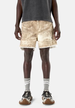 Mężczyzna noszący przetarte beżowe sztruksowe szorty, szare skarpetki za kostkę z białymi paskami oraz beżowo-czarne teksturowane sneakersy.