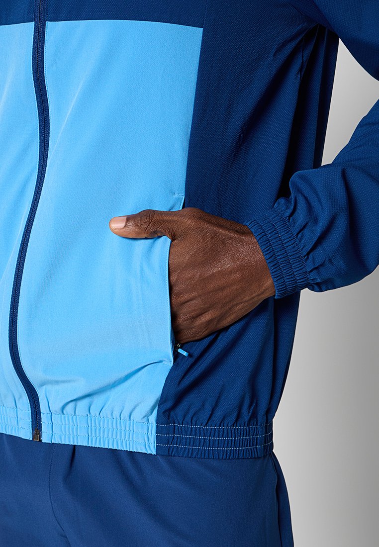 Mano nella tasca di una giacca sportiva zip-up blu e azzurra con polsini e vita elasticizzati, indossata con pantaloni blu navy.