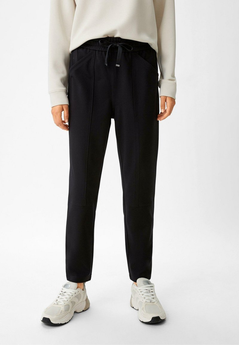 Black jogger pants with elastic waistband and drawstring, featuring side pockets and a streamlined design. Paired with light sneakers.