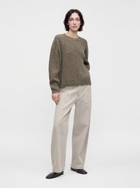 Femme portant un pull en maille vert olive, un pantalon beige à jambes larges avec de grandes poches, et des chaussures noires à bout carré, debout devant un fond blanc uni.
