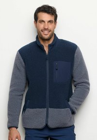 Giacca in pile navy e grigia con chiusura a zip, tessuto testurizzato, colletto alto, tasche laterali e taschino sul petto con zip.