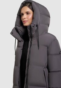 Chaqueta acolchada gris con cuello alto y capucha grande. Presenta un diseño acolchado, cierre frontal y cordones ajustables.