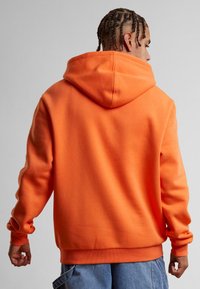 Oransje hettegenser med en avslappet passform, som har en stor hette, ribbestrikkede mansjetter og nederkant. Glatt stofftekstur, brukt over blå denimshorts.