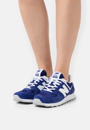Blaue Wildledersneaker mit weißen Akzenten, ausgestattet mit einer Netz-Zunge, flachen weißen Schnürsenkeln und einer Gummisohle. Auffälliges "N"-Logo an den Seiten.