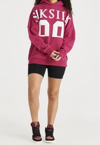 Persona vistiendo una sudadera oversized color marrón con texto blanco, pantalones cortos de ciclismo negros y zapatillas de caña alta negras y marrones, de pie contra un fondo blanco.