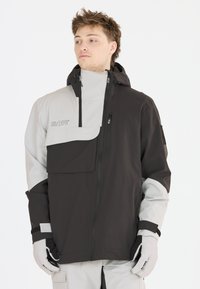 Veste de ski imperméable noire et grise avec un col haut, des poches zippées, une poche frontale à rabat et des poignets ajustables. Des gants blancs sont portés.