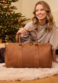 Rudas odinis sportinis krepšys su tvirtais rankenoms, matomais siūlais ir auksinėmis detalėmis, padėtas ant švelnaus balto kilimėlio.