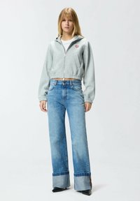 Felpa grigia corta con zip, caratterizzata da un logo a forma di cuore. Jeans azzurri a gamba larga con risvolto arrotolato, abbinati a stivali neri con tacco.