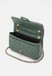 Borsa a mano verde in pelle con design trapuntato, hardware oro e tracolla a catena. Aperta, rivela un interno nero con una piccola tasca con zip.