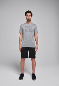 Grijze t-shirt met korte mouwen en een zwart geometrisch logo, gecombineerd met zwarte shorts. Model draagt zwarte sportschoenen. Eenvoudig, atletisch ontwerp.