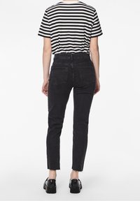 Persona vestida con vaqueros pitillo negros de tiro alto con dobladillo sin rematar, zapatos negros robustos y una camiseta de manga corta a rayas blancas y negras, de espaldas.