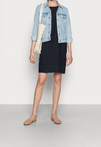 Osoba v světle modré džínsové bundě přes černé šaty, nese béžovou kabelku se vzorem přes rameno a má na sobě béžové sandály s otevřenou špičkou.