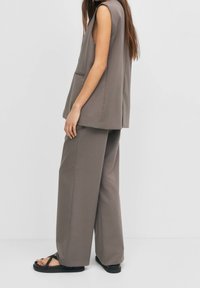 Kvinna i ärmlös taupe blazer, matchande vida byxor och svarta sandaler, stående på sidan mot en ljus, enfärgad bakgrund.