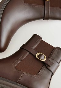 Bottes de cheville en cuir marron avec des panneaux élastiques, des accents de boucle dorée et une texture lisse. Design à bout rond et semelle robuste.