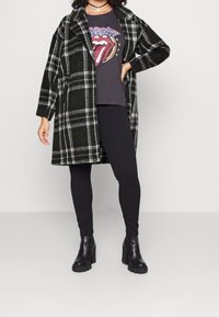 Zwart en groen geruit jasje met een losse pasvorm, gecombineerd met een grijze graphic t-shirt, zwarte leggings en chunky zwarte hakken.