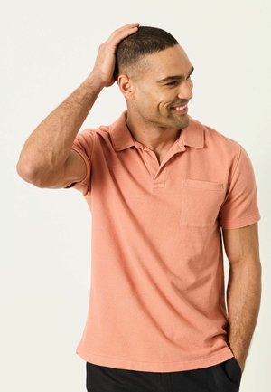 Mężczyzna z krótkimi włosami uśmiechający się, ubrany w łososiową koszulkę polo, dotykający głowy jedną ręką, stojący na tle jednolitego jasnego tła.