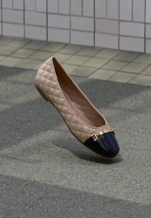 Chaussure plate matelassée beige avec bout noir et détail de boucle dorée posée sur un bitume gris près d’un mur carrelé.