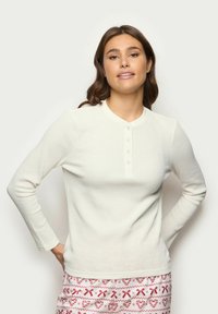 Maglietta henley bianca a maniche lunghe con una texture a coste, dotata di un colletto rotondo e quattro bottoni sul davanti.