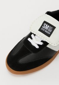 Schwarz-weißer Sneaker aus Wildleder und synthetischem Leder, mit weißen Schnürsenkeln, einem gepolsterten Kragen und einer beigefarbenen Gummisohle.