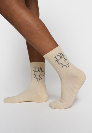 Benen met lichtbeige geribbelde sokken met een zwart verweerd tekstontwerp aan de zijkant, geplaatst tegen een effen lichte achtergrond.