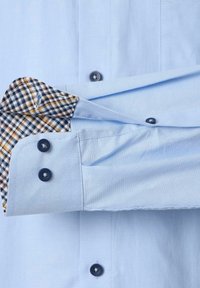 Lichtblauw shirt met donkere knopen en een geblokt stofaccent op de manchet. De manchet is netjes teruggevouwen, waardoor het patroon zichtbaar wordt.