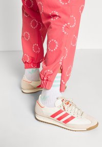 Gambe che indossano pantaloni Adidas color corallo con loghi circolari, sneaker Adidas bianche con strisce corallo e calze bianche con loghi trefoil Adidas verde chiaro.