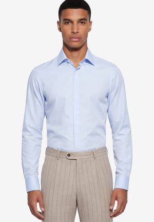 Mann trägt ein hellblaues Hemd, das in beige gestreifte Hose mit Einzelknopfverschluss gesteckt ist.