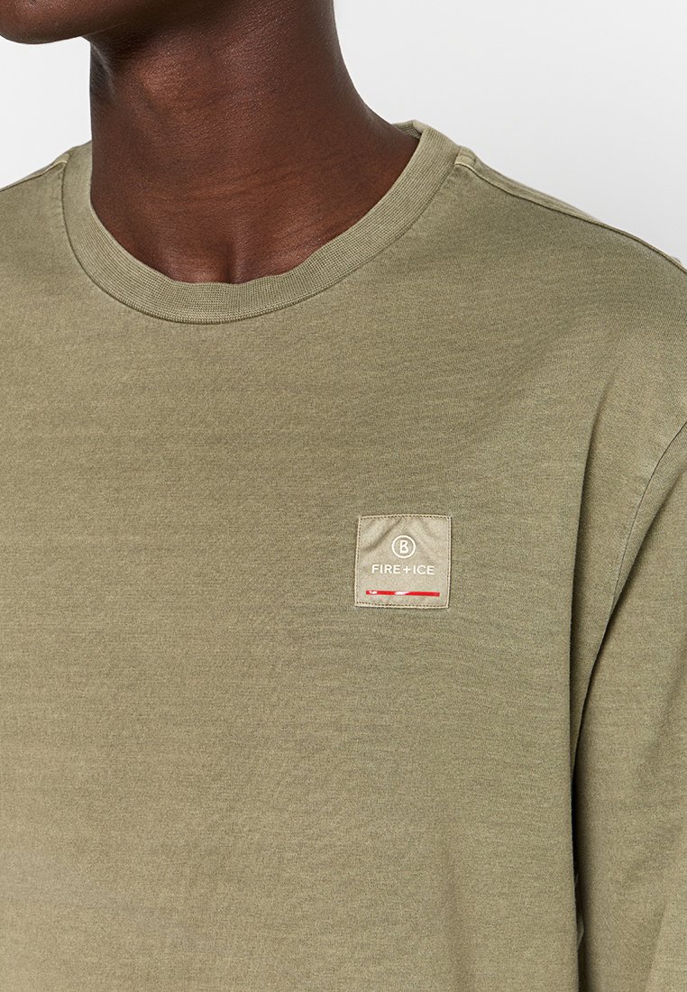Olivegrön bomulls-t-shirt med rund hals och en liten fyrkantig loggopatch med röda accenter på vänster bröst. Mjuk textur.