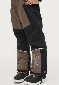 Bruine en zwarte skibroek met reflecterende accenten, met een geometrisch logo bij de knie en een duurzame, geïsoleerde stof.