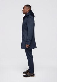Navy hooded coat with a textured finish, featuring a straight cut, side pockets, and button details, paired with dark trousers and casual shoes.
