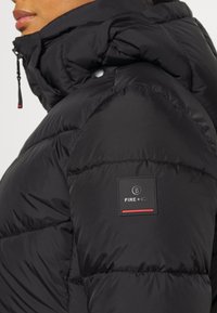Chaqueta acolchada negra con cuello alto, con detalles acolchados, un parche de logo en la manga y cierre frontal con cremallera.