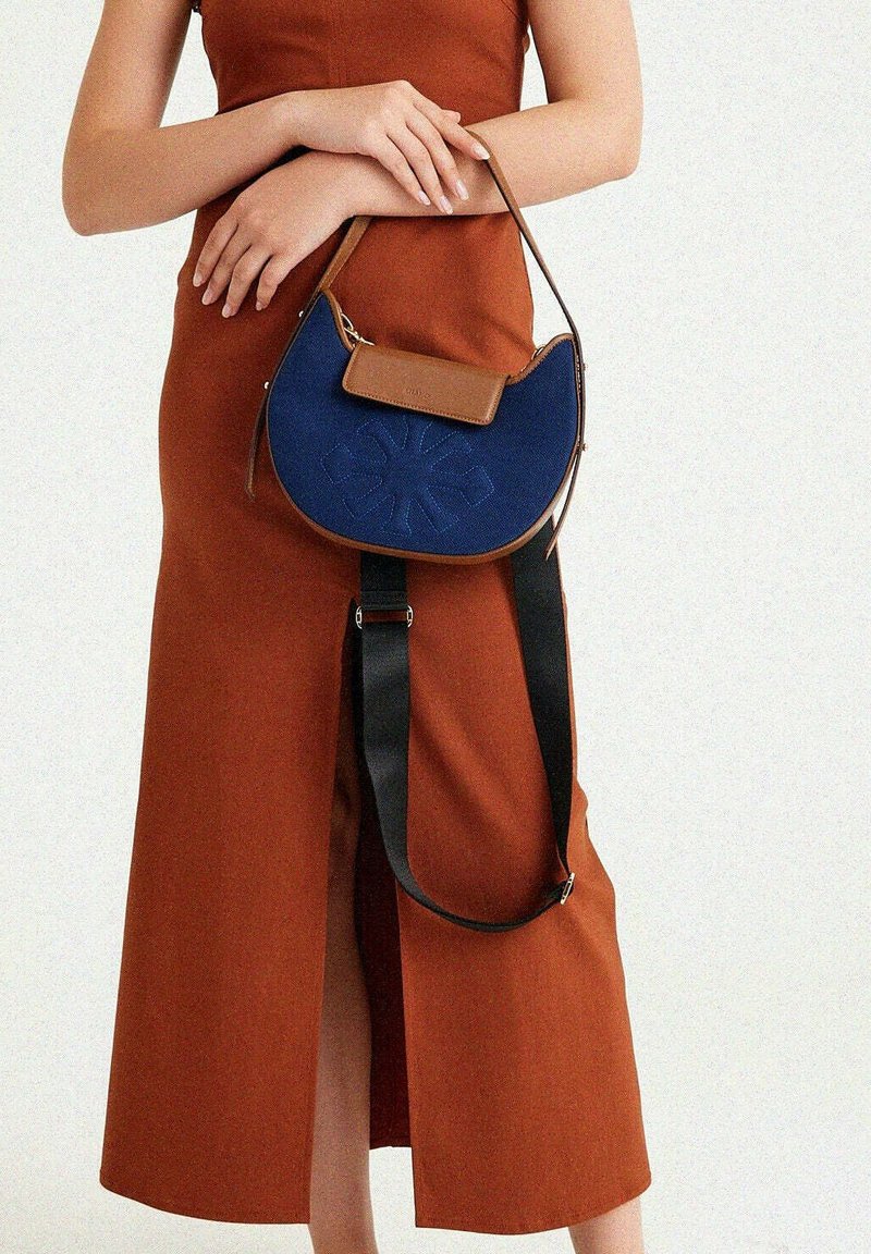 Blaue Lederhandtasche mit geschwungener Form, braunen Akzenten und einem strukturierten Blumenmuster. Verfügt über einen abnehmbaren schwarzen Tragegurt.