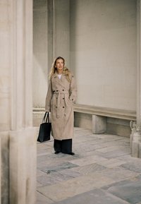 Kobieta w beżowym płaszczu i czarnych spodniach, trzymająca czarną torbę typu tote, stojąca w kamiennym korytarzu z kolumnami i kamienną ławką.