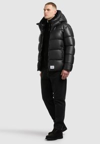 Homme portant une veste matelassée à capuche noire, un pantalon noir et des bottes noires, debout sur un fond gris clair.