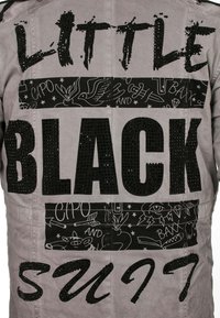 Graue Jacke mit auffälligem schwarzem Text "KLEINER SCHWARZER ANZUG", verziert mit schwarzen Strasssteinen. Die Muster umfassen Strichzeichnungen und stilisierte Grafiken.