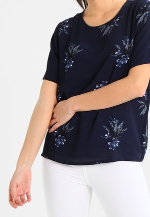 Donna che indossa una maglietta blu navy a maniche corte con motivi floreali blu e pantaloni bianchi, in piedi con una mano che tocca il braccio opposto.