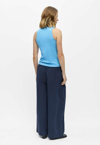 Mujer con cabello rubio hasta los hombros, que lleva una blusa azul sin mangas y pantalones oscuros de pata ancha, de pie de lado mirando a la derecha.