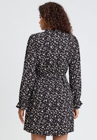 Robe noire à motifs floraux avec des fleurs blanches, manches longues avec des poignets volantés, taille nouée et texture de tissu lisse.