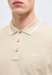 Gros plan sur une personne portant un polo beige avec un liseré blanc sur le col, trois boutons, et un logo "BOSS" sur la poitrine.