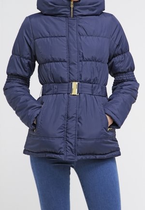 Granatowa pikowana kurtka puchowa z kapturem, zapinana na pasek, z zamykanymi na zamek kieszeniami, noszona z niebieskimi dżinsami, ręce w kieszeniach.