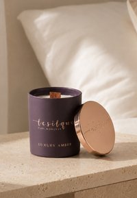 Bougie de luxe parfumée à l'ambre allumée dans un récipient en verre violet foncé avec une mèche en bois et un couvercle en cuivre posé à côté.